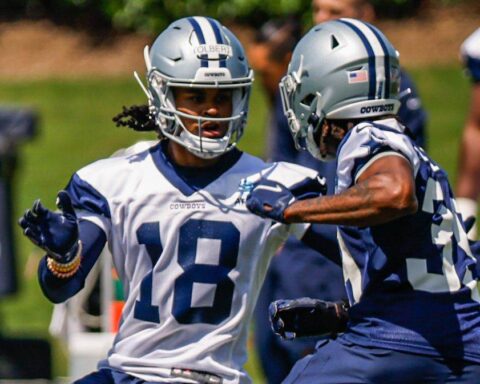 Two football players in Dallas Cowboys uniforms and helmets, including Jalen Tolbert, practice on the field with teammates in the background.