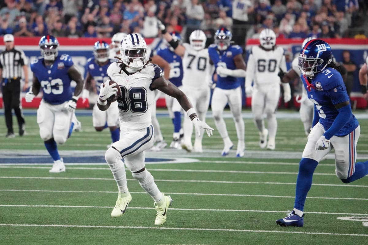 A football player in a white uniform runs with the ball while players in blue uniforms chase him on a football field.