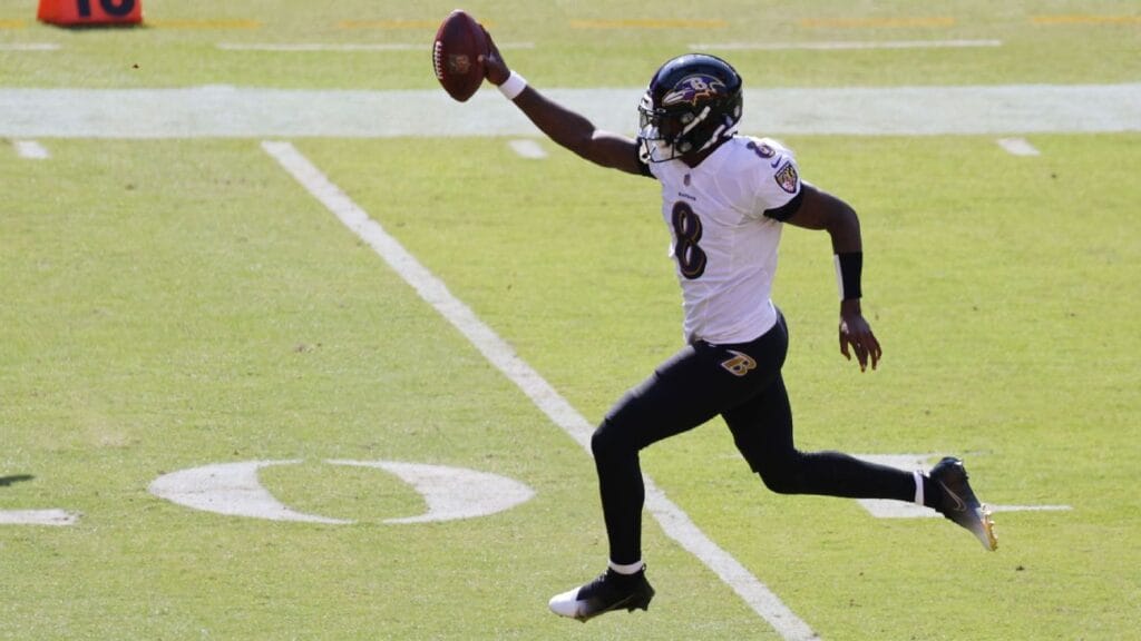 A football player in a white and black uniform runs with the ball on a green field, raising it above his head.