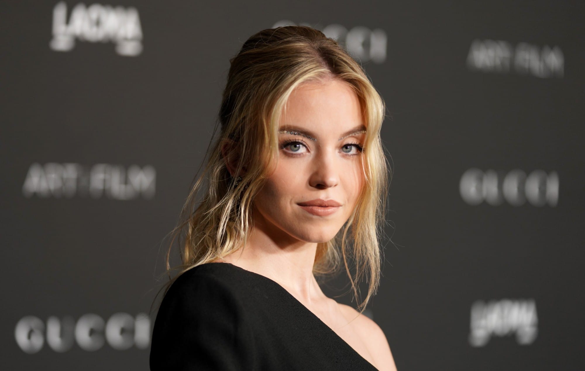 Person with blonde hair wearing a black off-shoulder outfit at an event with a dark backdrop.