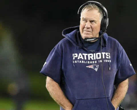 A football coach wearing a Patriots hoodie and headset stands on the field, looking off to the side.