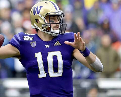 Quarterback in purple uniform preparing to throw a football on the field, with fans blurred in the background at a football game.