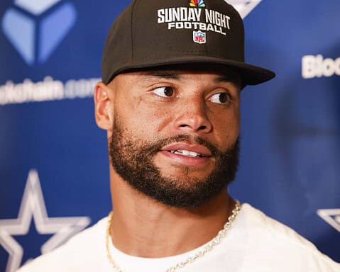NFL player wearing a black hat with "Sunday Night Football" logo and chains, speaking at a press conference, with Dallas Cowboys branding in the background.