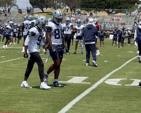 Dallas Cowboys football players during practice on the field, wearing team jerseys and helmets, with coaches and spectators in the background. Inside The Star.