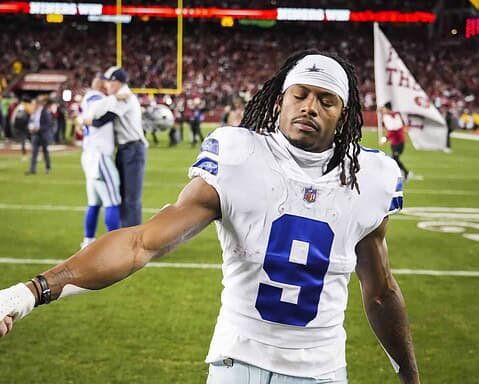 Helmetless Dallas Cowboys player on football field, holding hands with team member after game, showing sportsmanship and team unity, with cheering crowd in the background.