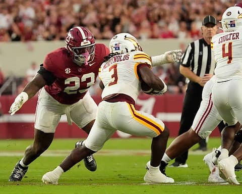 Alabama football linebacker #23 tackling a Kansas City Chiefs running back #3 during a game at a packed stadium.