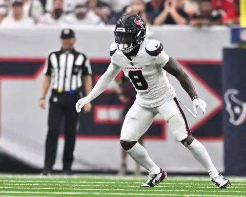 Houston Texans football player during game action, wearing white jersey number 8, on the field at NRG Stadium in Houston, Texas, with a blurred crowd and referee in the background.
