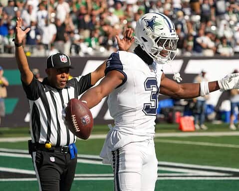 Dallas Cowboys football player celebrating after a touchdown, holding the football with an official behind him on the field during an NFL game.