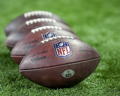 Close-up of four NFL footballs lined up on a green grass field, showcasing the official NFL logo, brand markings, and textured leather surface perfect for football game action pictures.