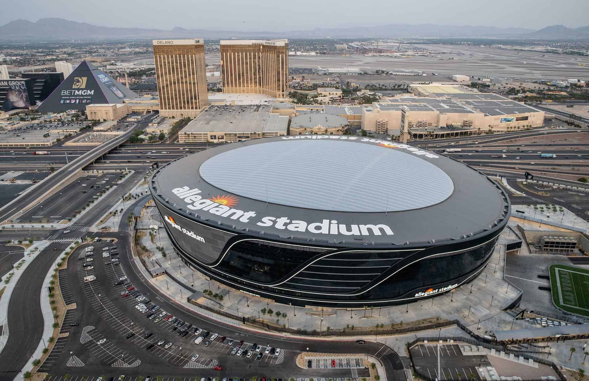 Sleek aerial view of Allegiant Stadium in Las Vegas, home to the Las Vegas Raiders NFL team, featuring its modern architecture, surrounding parking lots, and nearby city skyline.