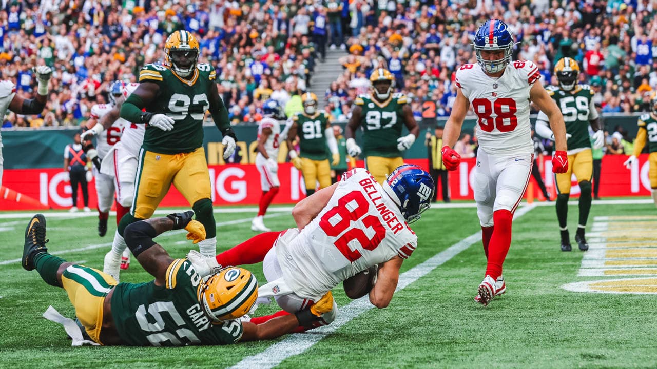 A Dallas Cowboys player with jersey number 88 is reaching for the end zone as he is tackled by a Green Bay Packers defender, number 55, during an exciting NFL game at Lambeau Field with a packed crowd cheering in the background.