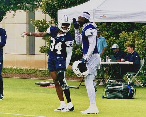 An image of two Dallas Cowboys football players in practice gear, one with a helmet and the other conversing, on a practice field with team staff and equipment in the background.