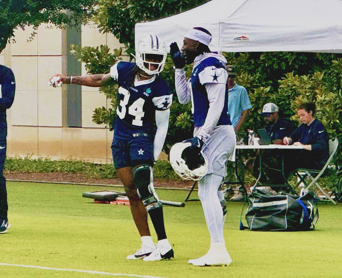 An image of two Dallas Cowboys football players in practice gear, one with a helmet and the other conversing, on a practice field with team staff and equipment in the background.