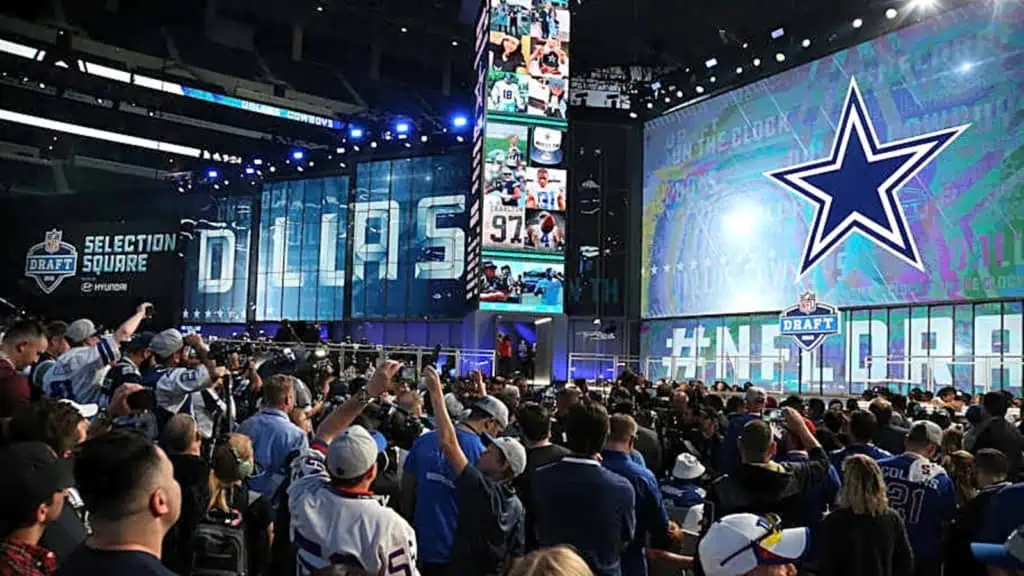 Draft day event with fans inside a stadium, showcasing large digital screens, team banners, and a vibrant atmosphere during the NFL Draft at the Cowboys' Selection Square.