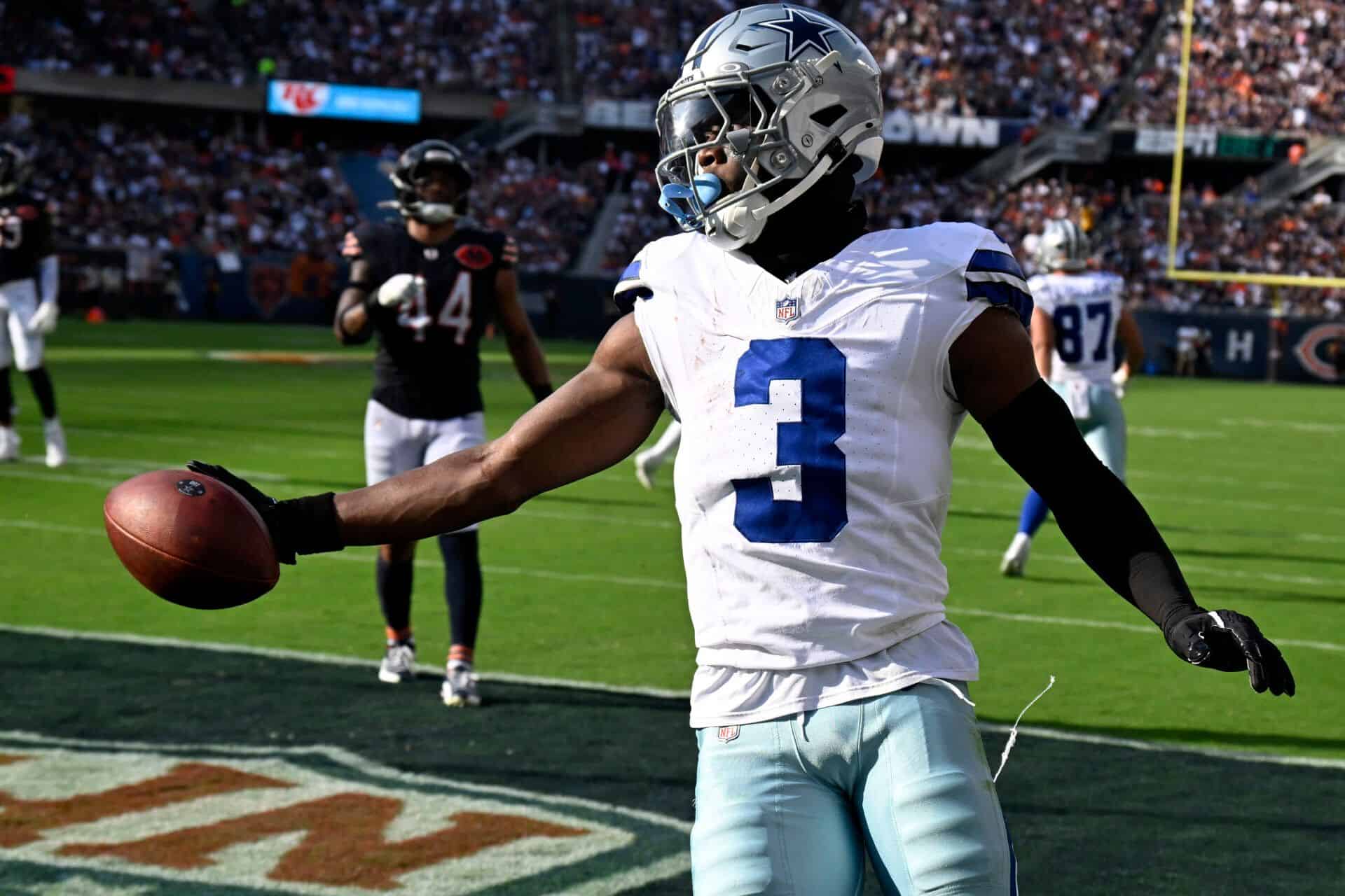 A professional football player from the Dallas Cowboys holding a football on the field during a game. The player is wearing a white jersey with the number 3, helmet, and black gloves, with other players and fans visible in the background.