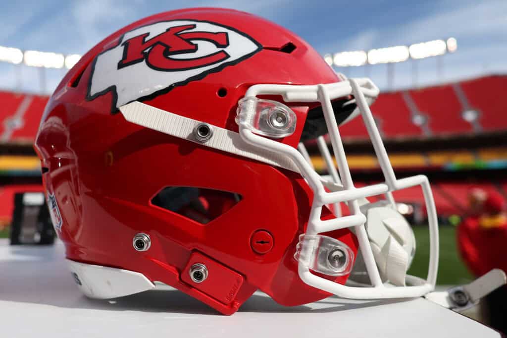 A close-up image of a red Kansas City Chiefs NFL helmet featuring the team logo and white facemask, displayed on a table at a football stadium during daytime.