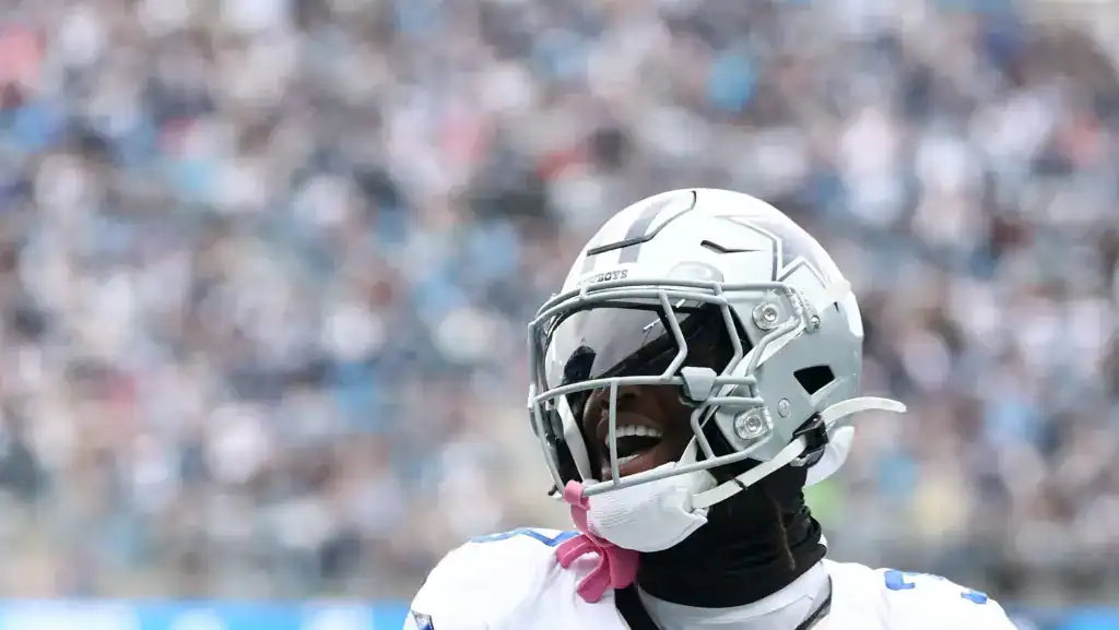 A close-up of a Dallas Cowboys football player in full gear, smiling and celebrating during a game, with a blurred stadium crowd behind him.