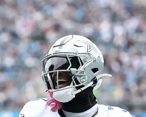 A close-up of a Dallas Cowboys football player in full gear, smiling and celebrating during a game, with a blurred stadium crowd behind him.