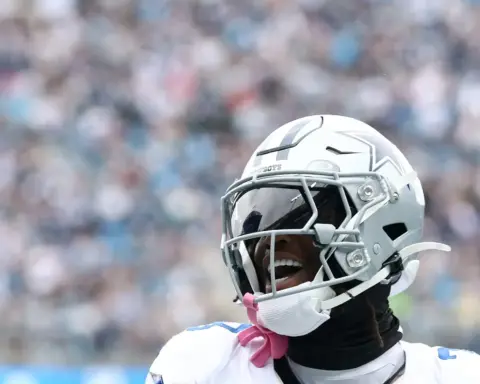 A close-up of a Dallas Cowboys football player in full gear, smiling and celebrating during a game, with a blurred stadium crowd behind him.