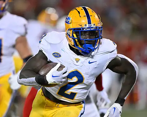 A college football player from the University of Georgia (UGA) in yellow and white uniform carries the football during a game, showcasing athleticism and team spirit, with other players and the stadium background.