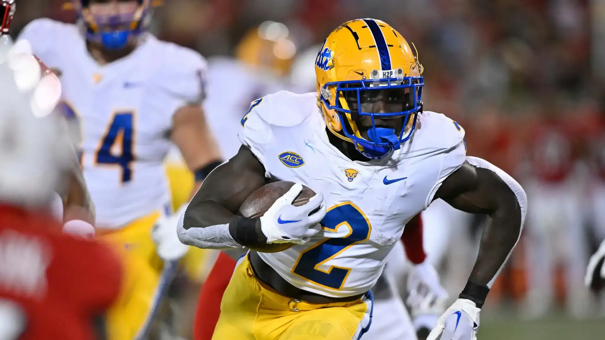A college football player from the University of Georgia (UGA) in yellow and white uniform carries the football during a game, showcasing athleticism and team spirit, with other players and the stadium background.
