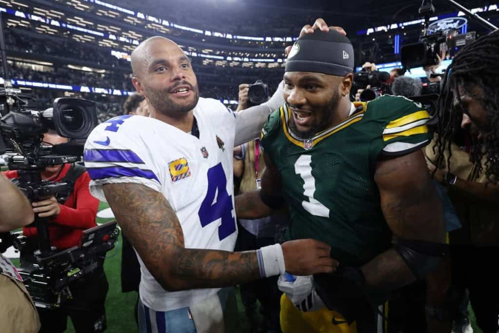 Celebrating victory, Dallas Cowboys and Green Bay Packers players share a moment on the field after an NFL game, surrounded by cameras and media at the stadium.
