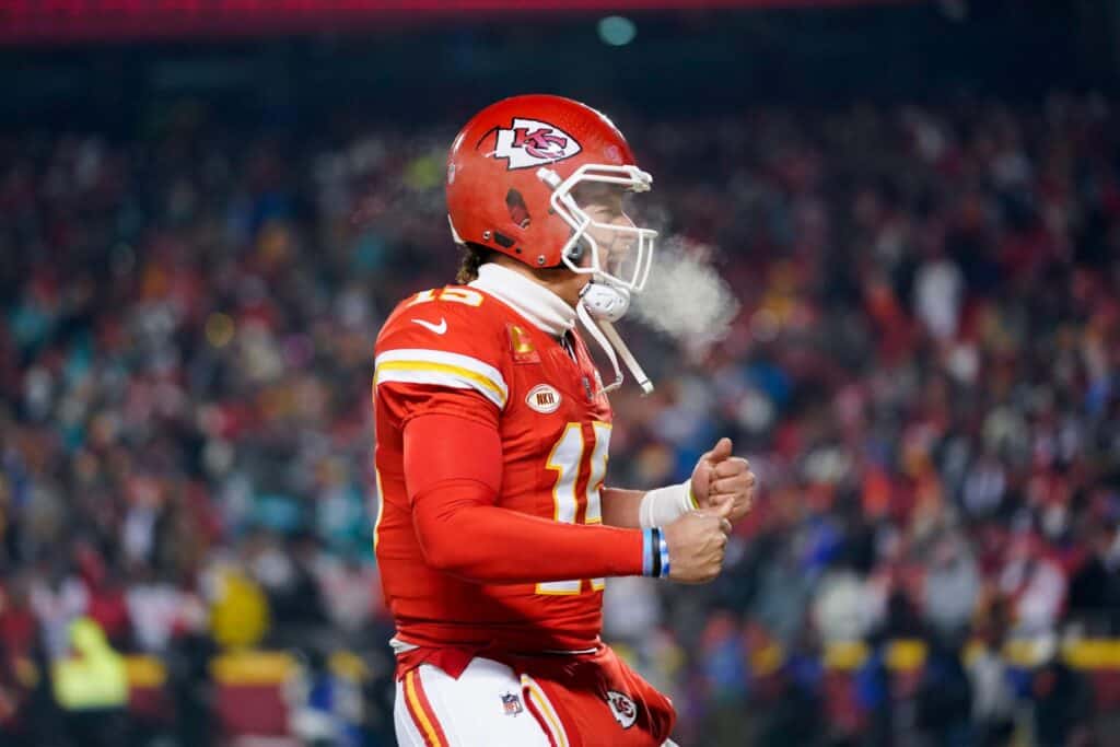 Kansas City Chiefs football player wearing helmet and uniform, celebrating on the field during a game, with fans in the background.