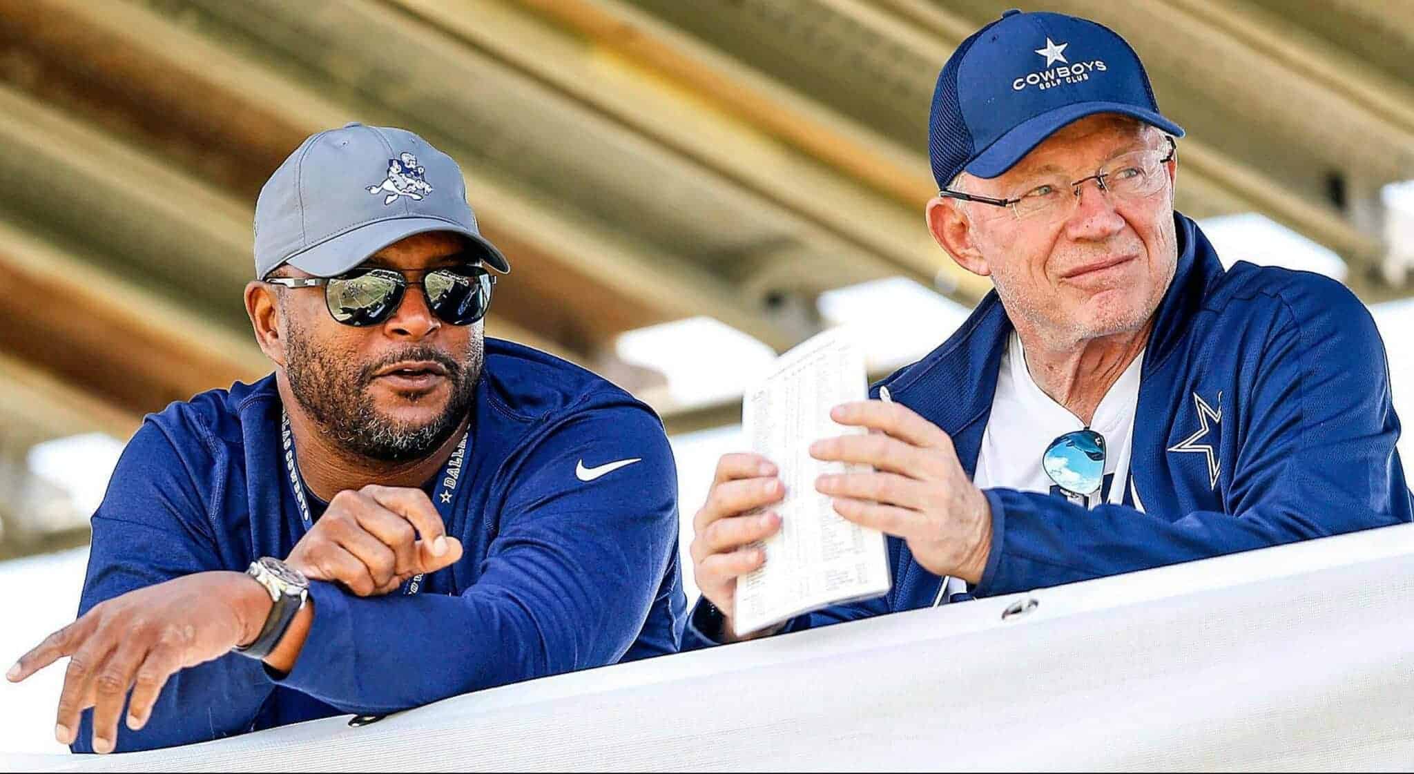 Inside The Star Dallas Cowboys football coaches discussing strategies outdoors in team gear, wearing caps, sunglasses, and jackets with the Dallas Cowboys logo during a game or practice.