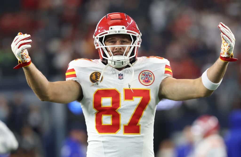 A football player in a white Kansas City Chiefs jersey and red helmet with outstretched arms, making a celebratory gesture on the field during a competitive NFL game.