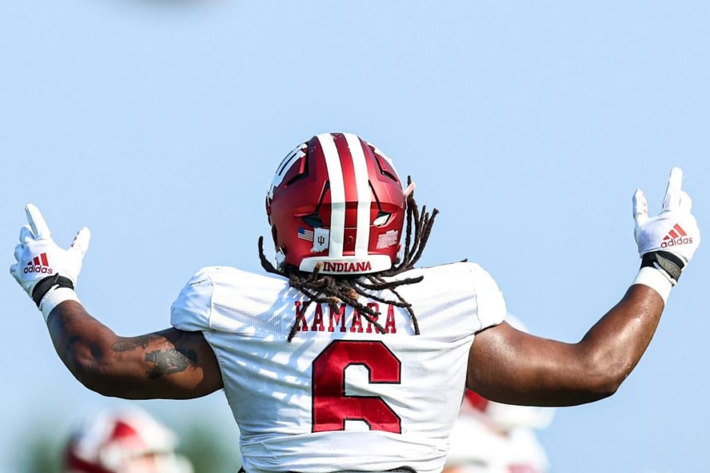 Indiana Hoosiers football player, Mikail Kamara, wearing a white No. 6 jersey, raises both arms in celebration while facing away from the camera during a daytime game