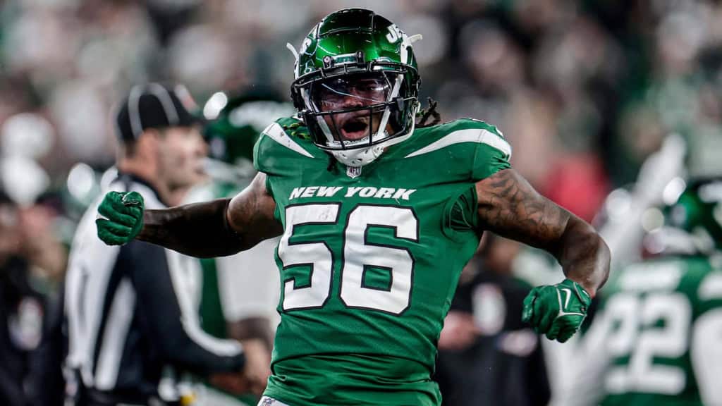 New York Jets linebacker wearing a green number 56 jersey celebrates on the field with arms flexed during a night game, with the crowd blurred in the background.