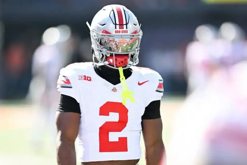 Ohio State Buckeyes defensive back, Caleb Downs, in a white No. 2 jersey and silver helmet with a mirrored visor stands on the field during warmups, wearing a neon green mouthguard, with the stadium background blurred.