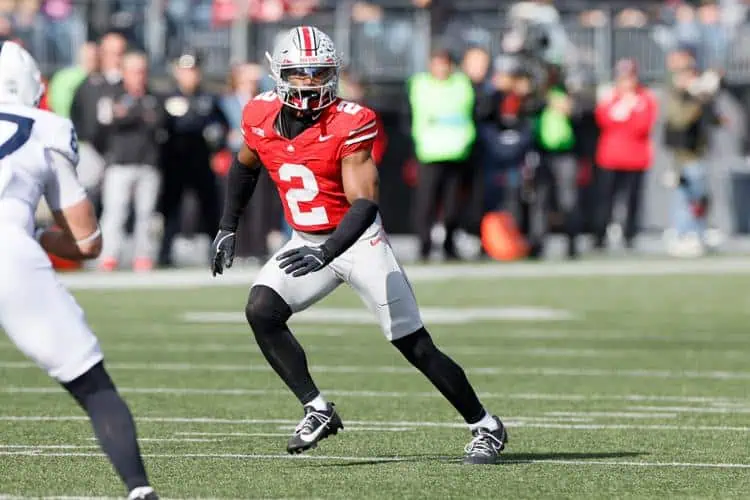 Ohio State Buckeyes defensive back, Caleb Downs, wearing a red No. 2 jersey and silver helmet lines up in coverage on the field during live game action, knees bent and eyes forward, with opposing players and spectators blurred in the background.