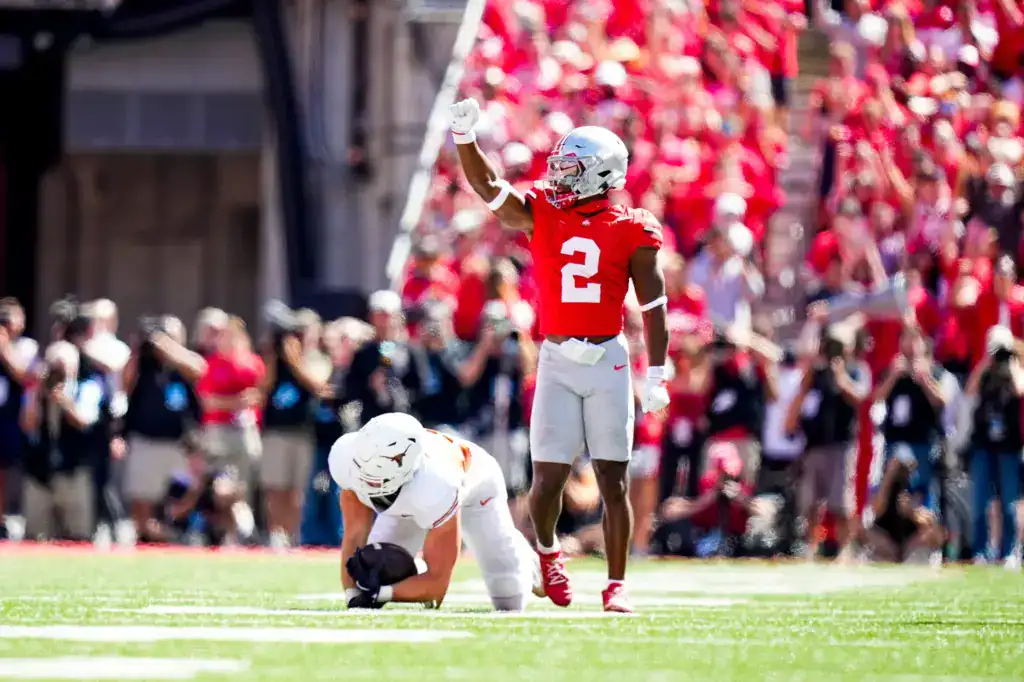 Ohio State Buckeyes defensive back, Caleb Downs, in a red No. 2 jersey raises his arm to signal fourth down as an opposing player kneels on the turf, with a packed crowd in the background.