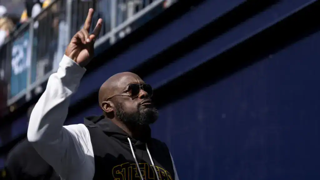 Pittsburgh Steelers head coach Mike Tomlin, wearing sunglasses and a black Steelers hoodie raises a two-finger gesture while walking along the sideline of an NFL stadium before a game. 