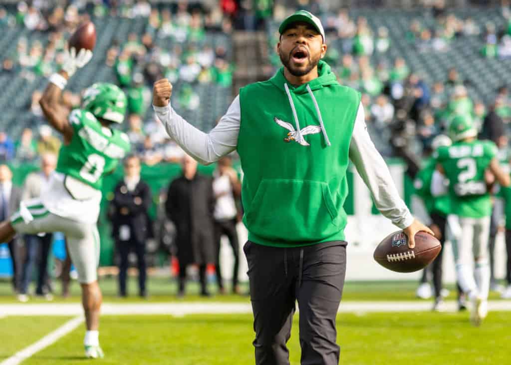 A Philadelphia Eagles coach Christian Parker wearing a green sleeveless hoodie and black pants walks on the field during warmups, holding a football and raising his fist as players in green uniforms warm up behind him.