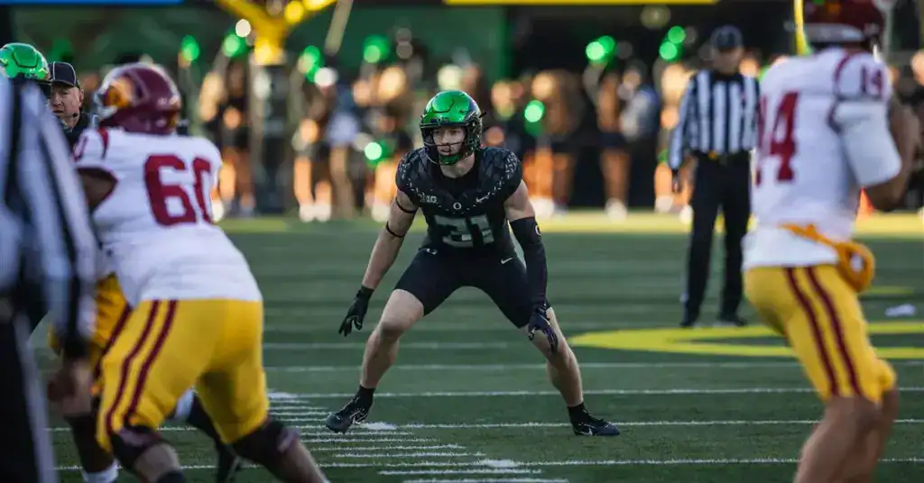 Oregon Ducks safety, Dillon Thieneman, in a black uniform and green helmet lines up in a defensive stance before the snap against USC, with opposing players in white and red uniforms at the line of scrimmage.