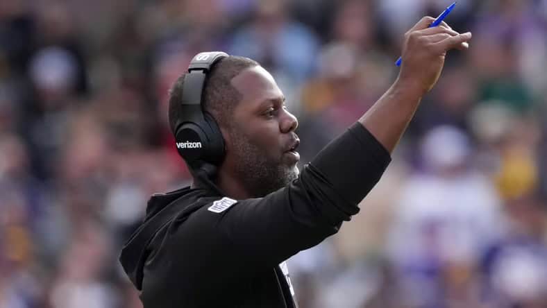 Minnesota Vikings coach, Daronte Jones, stands on the sideline  with his arm in the air, in front of a blurred image of the crown in the stands.