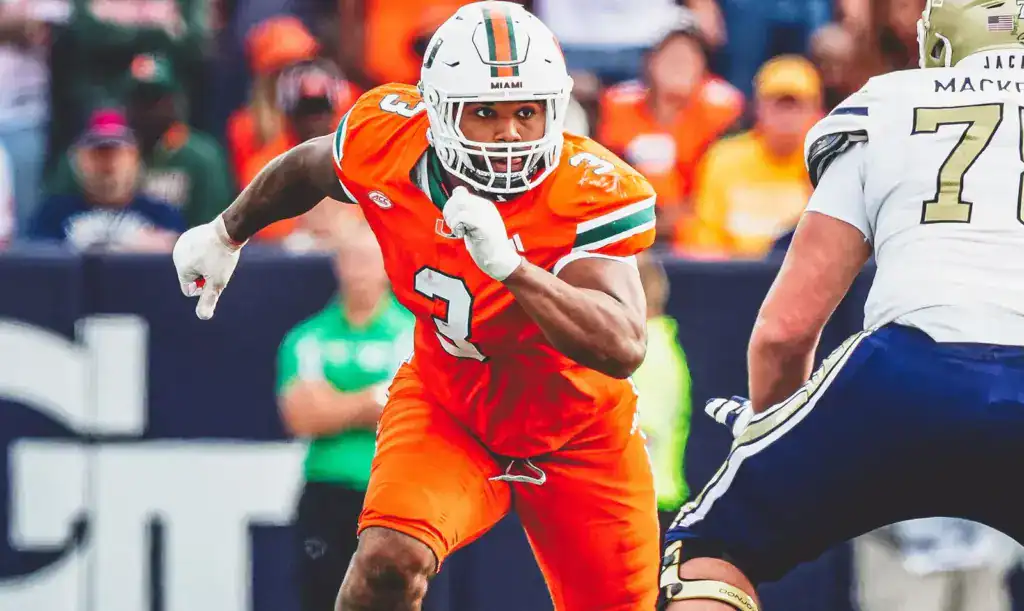 Miami Hurricanes defensive lineman, Akheem Mesidor, in an orange uniform sprinting with the football as an offensive lineman closes in, showing intense focus during a game play.