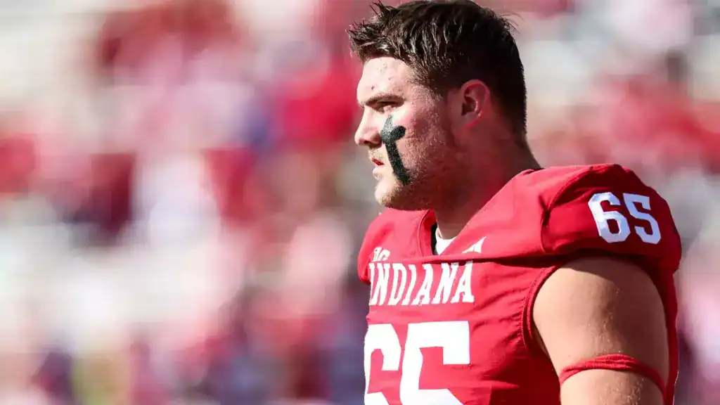 Indiana football offensive lineman, Carter Smith, wearing a red jersey with number 65, black eye paint on his cheek, standing on the sideline and looking toward the field during a game.