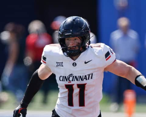 Cincinnati Bearcats linebacker, Jake Golday, in a white No. 11 uniform runs forward with arms extended during a daytime college football game.