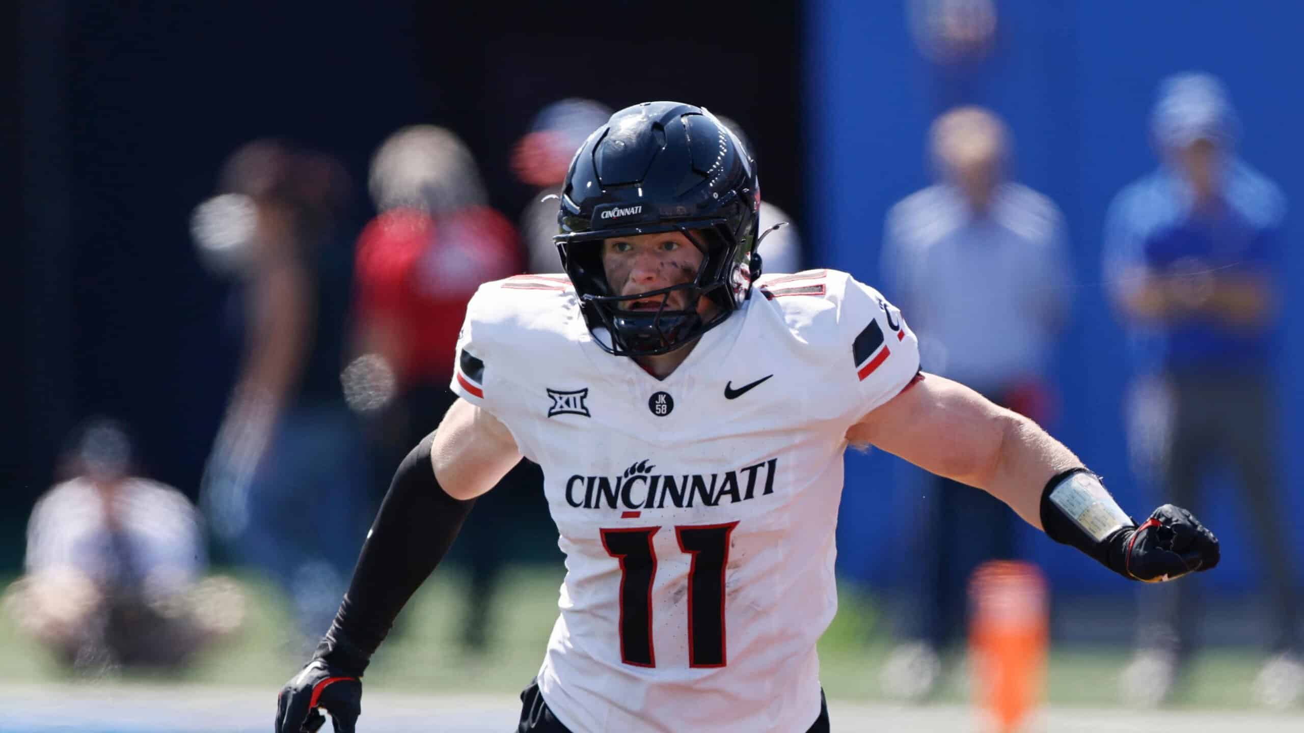 Cincinnati Bearcats linebacker, Jake Golday, in a white No. 11 uniform runs forward with arms extended during a daytime college football game.