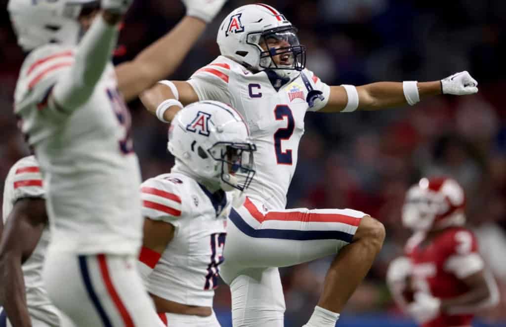 Arizona Wildcats football player wearing a white No. 2 jersey leaps and celebrates with arms extended while teammates react around him during a night game.