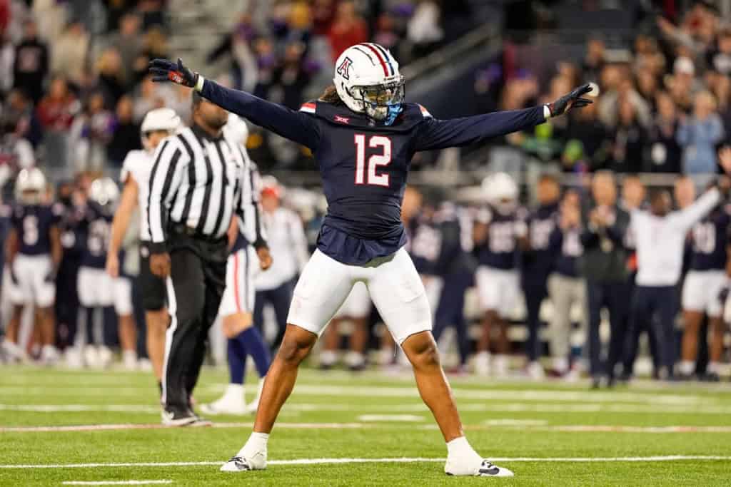 Arizona Safeties the Cowboys Should Target in 2026 NFL Draft NFL Draft Prospect and Arizona Wildcats football player, Genesis Smith, wearing a navy No. 12 jersey stands on the field with arms extended wide in celebration during a night game as teammates and officials look on.