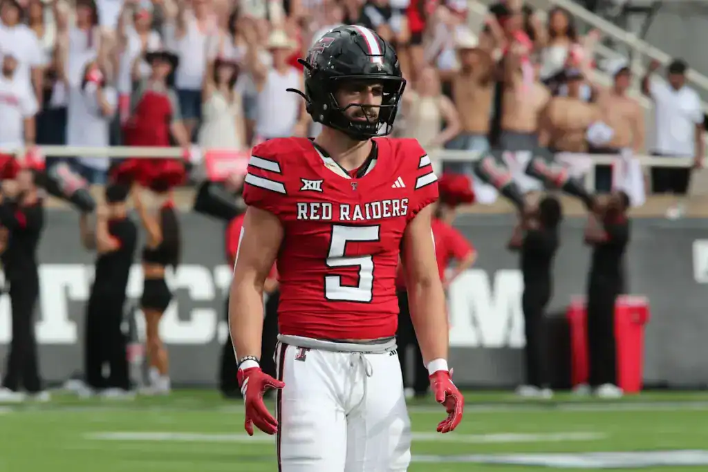 Texas Tech Red Raiders safety, Cole Wisniewski, wearing a red No. 5 jersey stands on the field during a daytime game as fans cheer in the stands behind him.