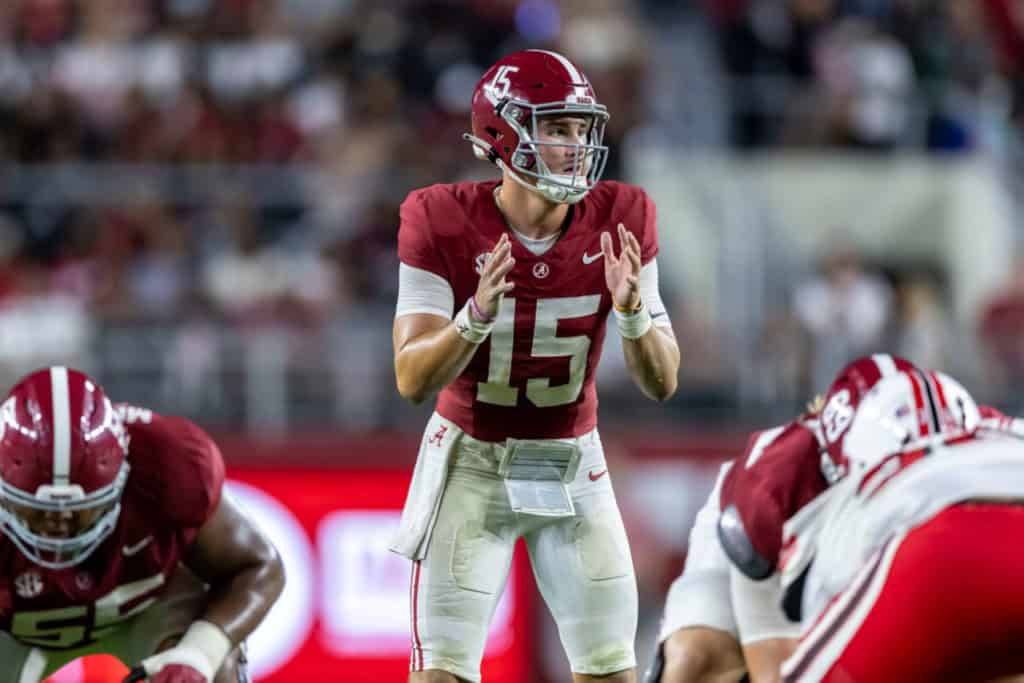 Alabama Crimson Tide quarterback, Ty Simpson, wearing a crimson No. 15 jersey stands at the line of scrimmage with hands raised, calling signals before the snap as offensive linemen set around him during a night game, capturing the hype and intensity of a big-time college football moment.