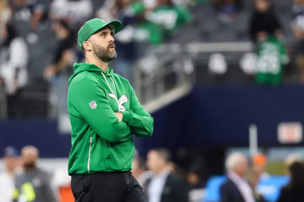 Philadelphia Eagles coach, Nick Sirianni, in a green hoodie and cap stands on the sideline with arms crossed, watching the field with a focused expression during an NFL game.