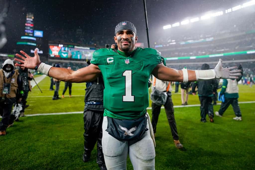 Philadelphia Eagles quarterback wearing a green No. 1 jersey celebrates on the field with arms spread wide after a game, with stadium lights, rain, and photographers surrounding him.