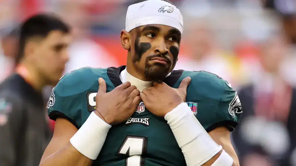 Philadelphia Eagles quarterback in a green uniform and white headband adjusts his jersey on the sideline, showing a serious, locked-in expression before kickoff.
