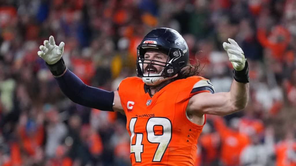 Denver Broncos soon-to-be free agent defensive player in an orange No. 49 jersey raises both arms in celebration during an NFL game, showing intensity and energy in front of a loud home crowd.